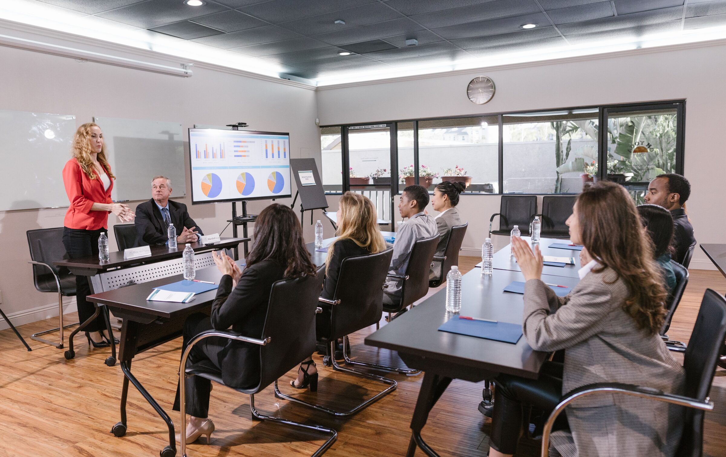 A presenter addresses a seated group gathered around a conference table, who are listening closely.