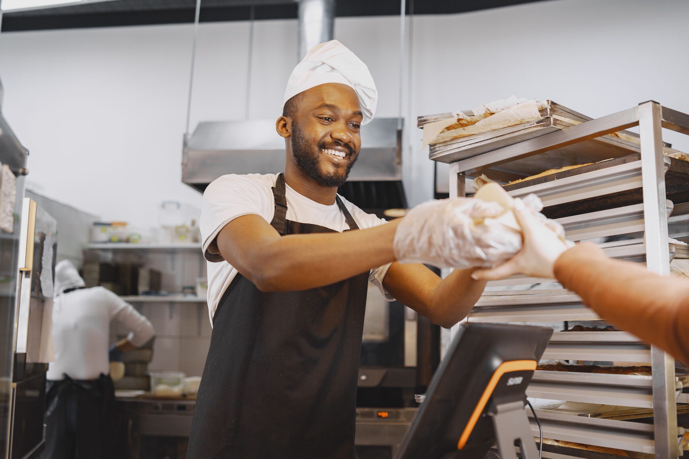 A baker hands off a tray of dough to a coworker in a commercial kitchen, both focused on the work.