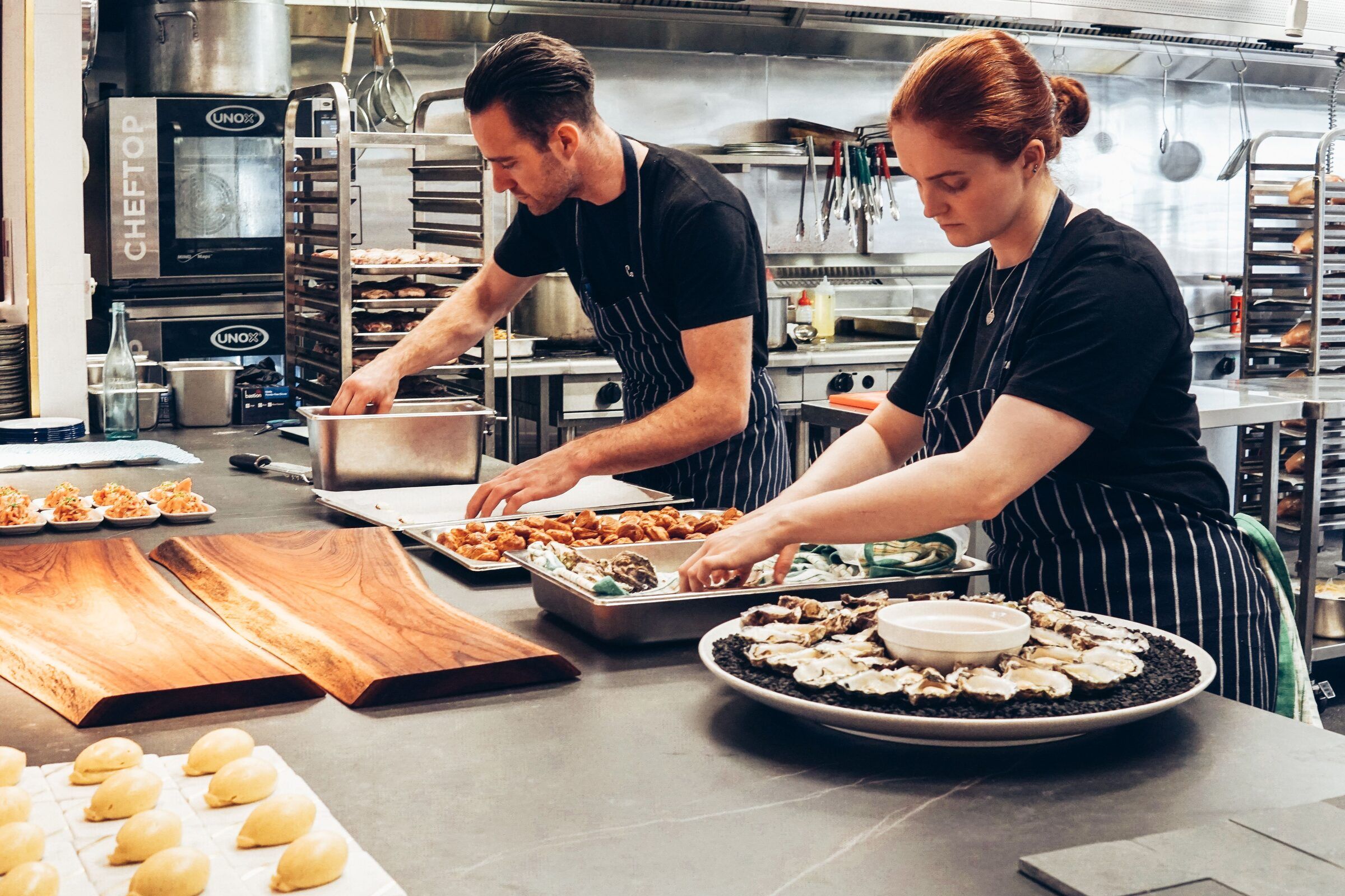 Two chefs in a restaurant kitchen plate oysters and prepared dishes, working side by side.